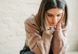 Crop unhappy female in casual wear touching cheeks and looking down while sitting near white wall in light room at home