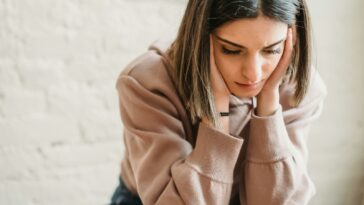 Crop unhappy female in casual wear touching cheeks and looking down while sitting near white wall in light room at home