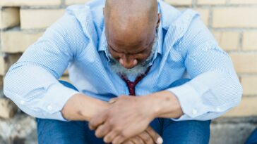 Hairless senior ethnic male in trendy outfit sitting near street wall and looking down in crisis
