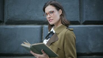 From below confident young female in glasses and elegant vintage clothes looking away and contemplating while reading rook on street near black wall of aged building in city