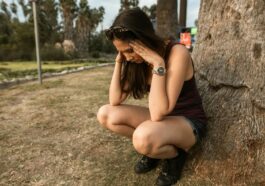 A woman kneels by a tree holding her head, expressing stress or anxiety in an outdoor park.