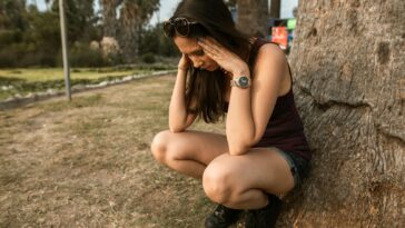 A woman kneels by a tree holding her head, expressing stress or anxiety in an outdoor park.