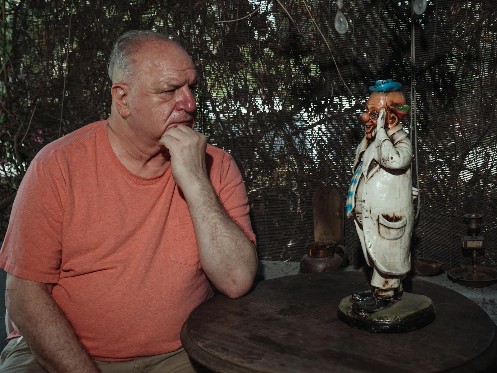 An elderly man in deep thought sits next to a whimsical statue outdoors.