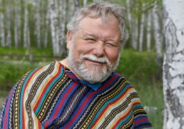 A cheerful senior adult man in a colorful poncho smiling amidst birch trees in a forest setting.