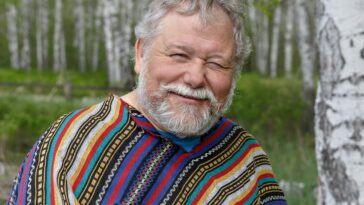 A cheerful senior adult man in a colorful poncho smiling amidst birch trees in a forest setting.