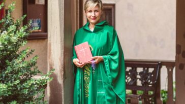 A woman clergy wearing a green robe, holding a Bible and rosary on an outdoor porch.