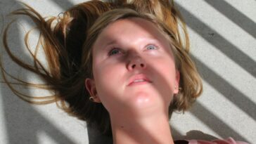 Close-up of a young woman lying on a floor with dramatic shadows and sunlight casting across her face.