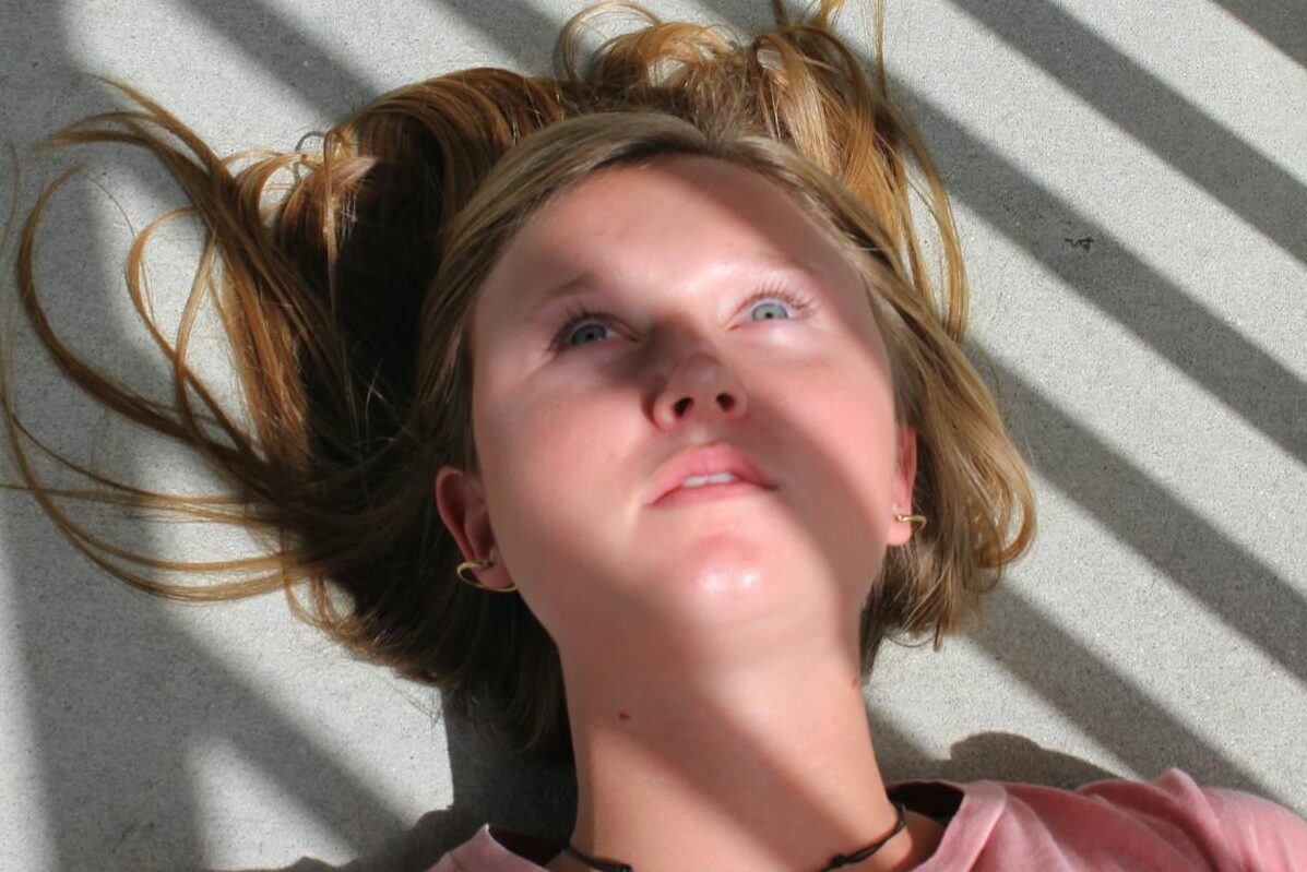 Close-up of a young woman lying on a floor with dramatic shadows and sunlight casting across her face.