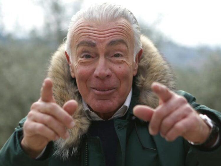 A joyful elderly man in a green parka smiling and pointing outdoors on a clear day.