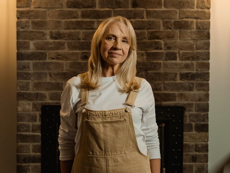 A senior woman in overalls smiles against a brick wall, warmly lit indoors.