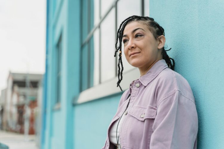 Ethnic middle aged female with braided hairstyle looking into distance while standing near wall of residential blue house in town