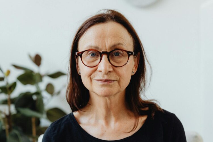 A close-up portrait of a mature woman indoors with eyeglasses, natural lighting.