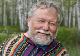 A cheerful senior adult man in a colorful poncho smiling amidst birch trees in a forest setting.
