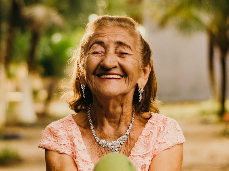Smiling elderly woman with jewelry showcases happiness while holding a mango outdoors.