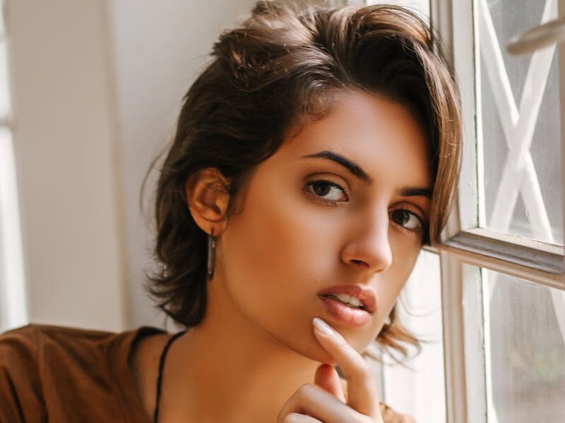 Graceful portrait of a thoughtful woman leaning against a window, capturing natural beauty and introspection.
