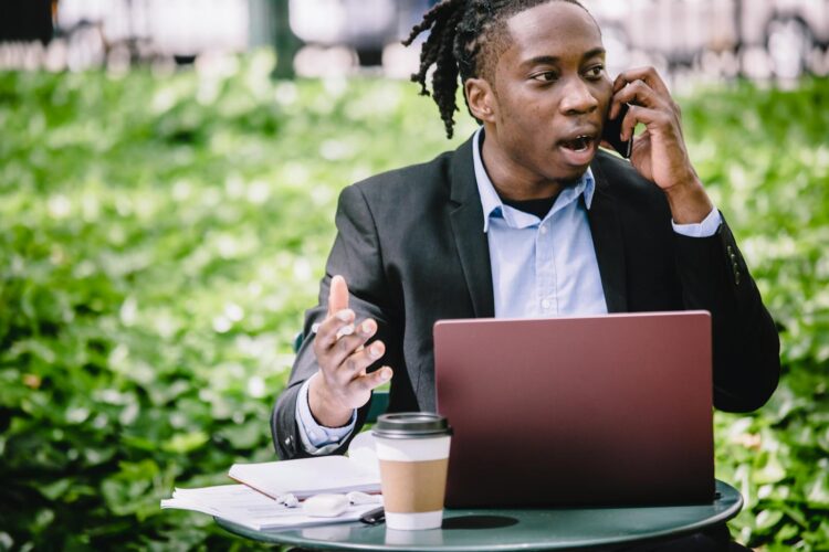 Young African American male entrepreneur in smart suit sitting in outdoor cafe with laptop and arguing on cellphone on sunny day