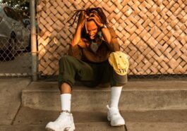 A man sits pensively on a curb, hands in hair, in a warm urban environment.
