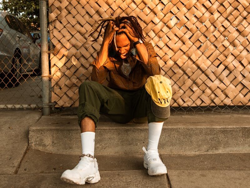 A man sits pensively on a curb, hands in hair, in a warm urban environment.