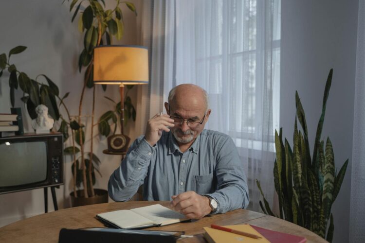 A senior man with eyeglasses works at a wooden table in a cozy home office.