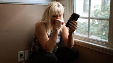 Woman sitting in deep thought by a window, holding a smartphone.