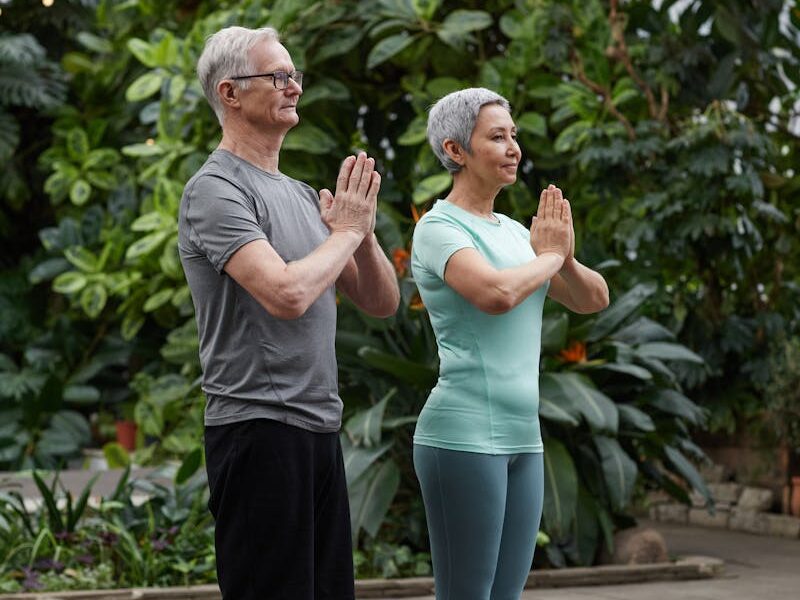 Senior couple practicing yoga poses together in a lush greenhouse setting.