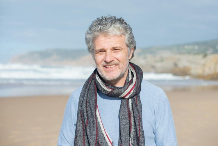 A senior man with gray hair and a scarf smiling on a sunny beach in Portugal.