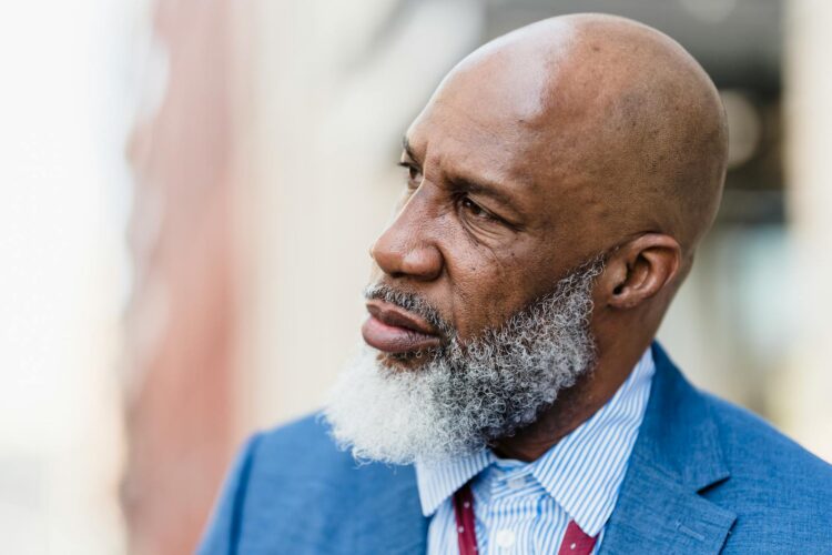 Elderly man with a white beard in a blue suit thoughtfully gazing outdoors.