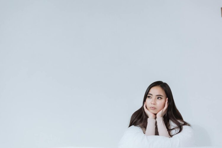 A thoughtful Asian woman gazes pensively with hands on chin in a minimalist studio.