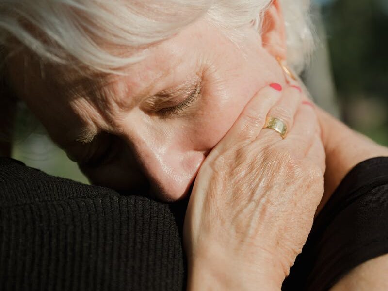 Elderly woman with white hair hugging a person, expressing emotion and connection.