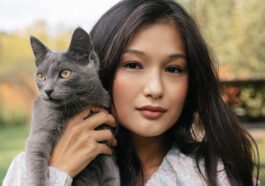 A young woman holds a gray cat outdoors in a serene setting, showcasing companionship and beauty.