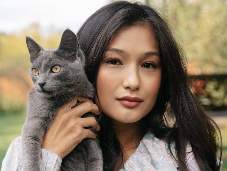 A young woman holds a gray cat outdoors in a serene setting, showcasing companionship and beauty.