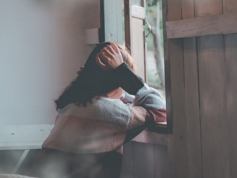 A woman sits indoors gazing thoughtfully out of a window, evoking emotions of loneliness and reflection.