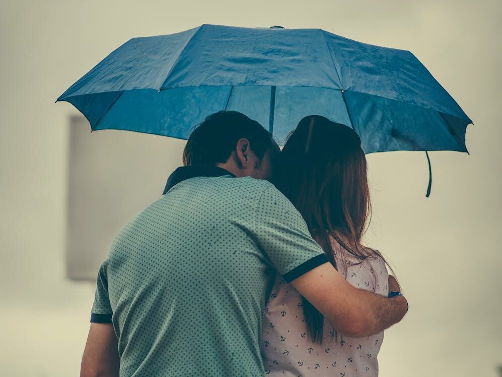 A couple embraces under a blue umbrella in a rainy street scene, evoking love and warmth.