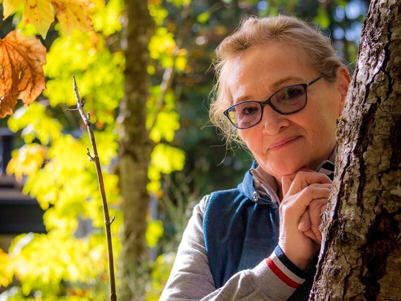 Smiling senior woman with glasses enjoys a sunny autumn day outdoors near a tree.