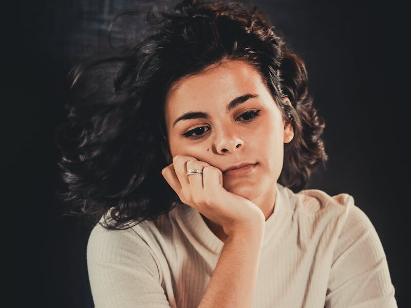 Portrait of a woman sitting pensively, wearing a casual white top. Moody, introspective atmosphere.