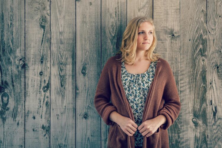 A woman stands by a rustic wooden wall, looking contemplative in casual attire.