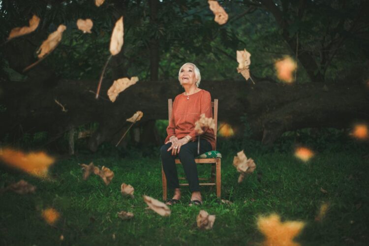 An elderly woman sits outdoors enjoying the falling autumn leaves, capturing a serene moment.