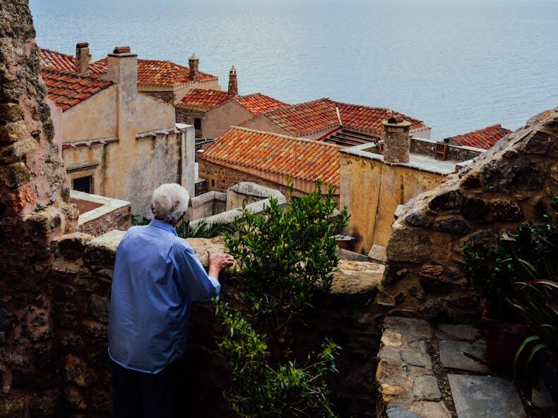 An elderly man overlooks a scenic coastal town with red rooftops and a vast blue sea under a cloudy sky.