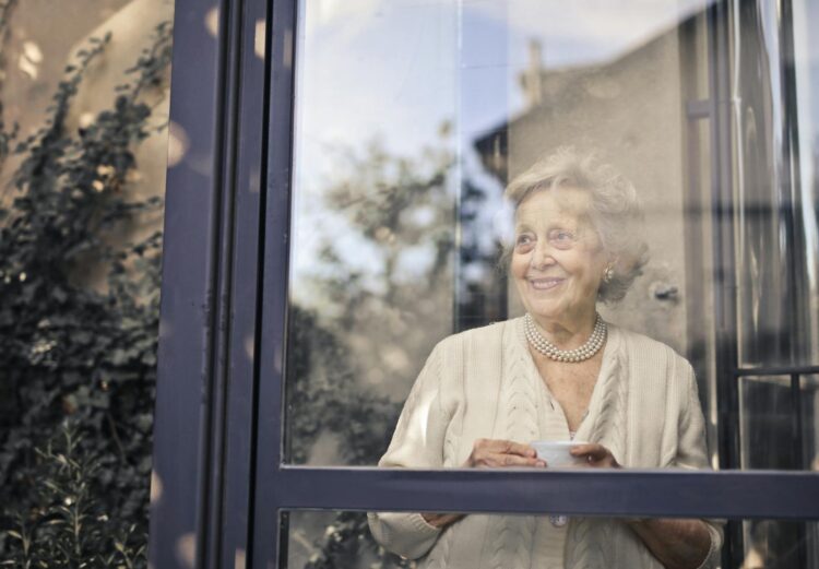 Elderly woman smiles by a glass window, holding a cup, in a peaceful setting.