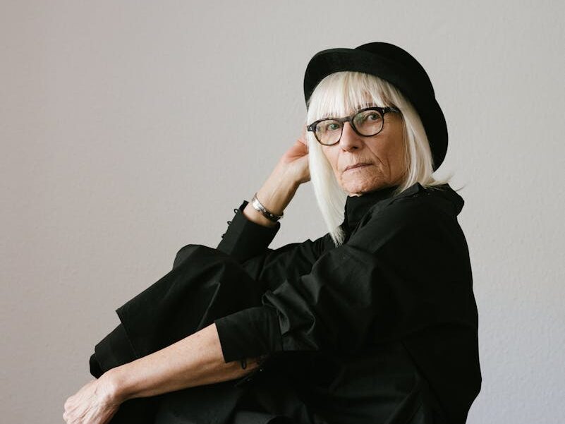 Elegant senior woman wearing eyeglasses and a black outfit posing indoors, showcasing timeless fashion.