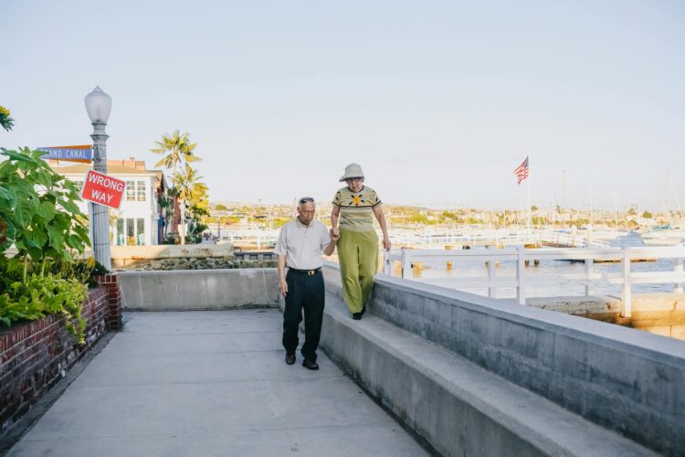 A senior couple walks hand in hand by a peaceful marina, enjoying a sunny day outdoors.
