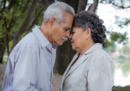 An elderly couple lovingly embraces with heads touching in a serene outdoor setting.