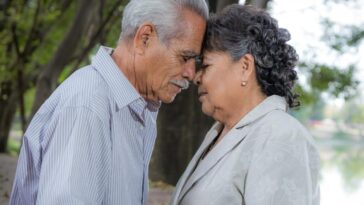 An elderly couple lovingly embraces with heads touching in a serene outdoor setting.