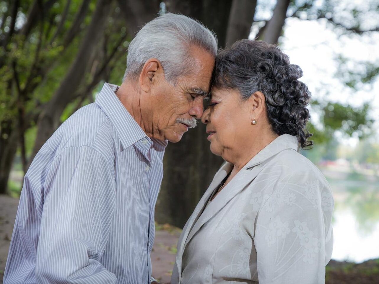 An elderly couple lovingly embraces with heads touching in a serene outdoor setting.