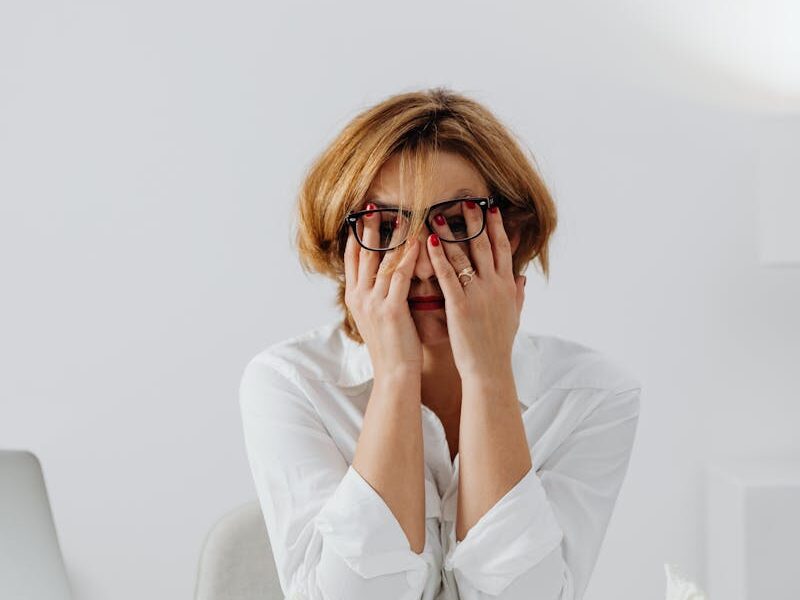 A stressed woman in eyeglasses and white shirt sits at her desk, overwhelmed and exhausted.