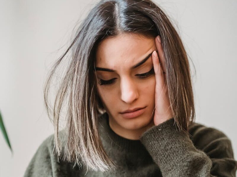 Unhappy female in casual wear leaning on hand and surfing internet on cellphone while sitting in light room near wall at home
