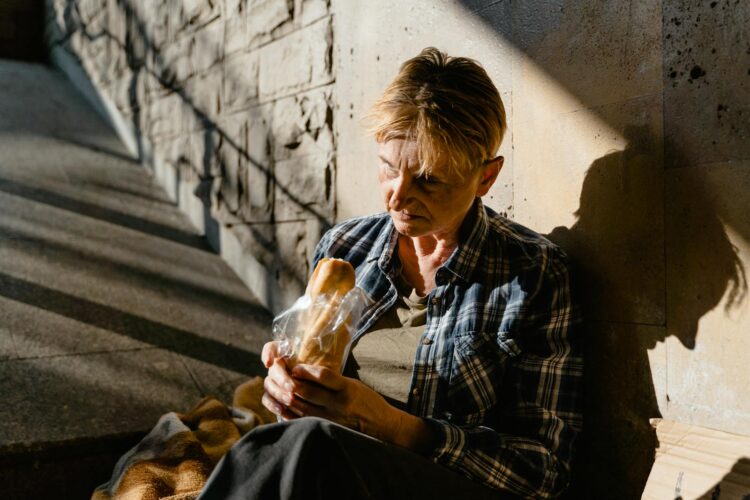 A senior woman holding and eating bread while seated against a sunlit wall, casting a thoughtful shadow.