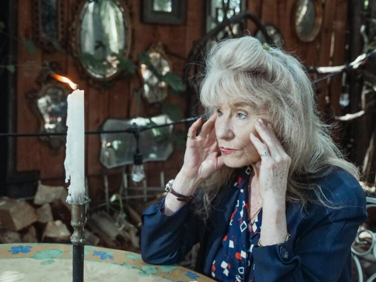 A thoughtful senior woman sits by a candle in an intimate, mirror-lined interior setting.