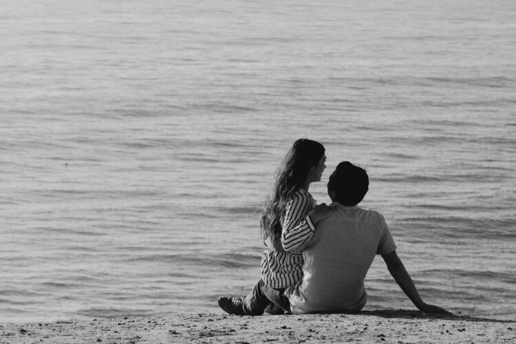 Black and white photo of a couple sitting close by the ocean, depicting love and tranquility.
