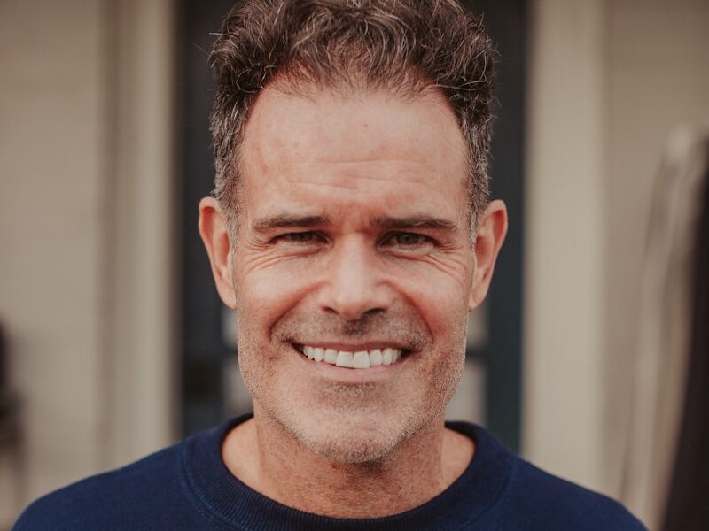 A smiling man in a blue sweatshirt stands in front of a door